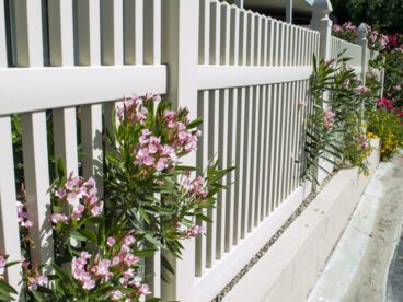 White vinyl fence with plants poking through over sidewalk