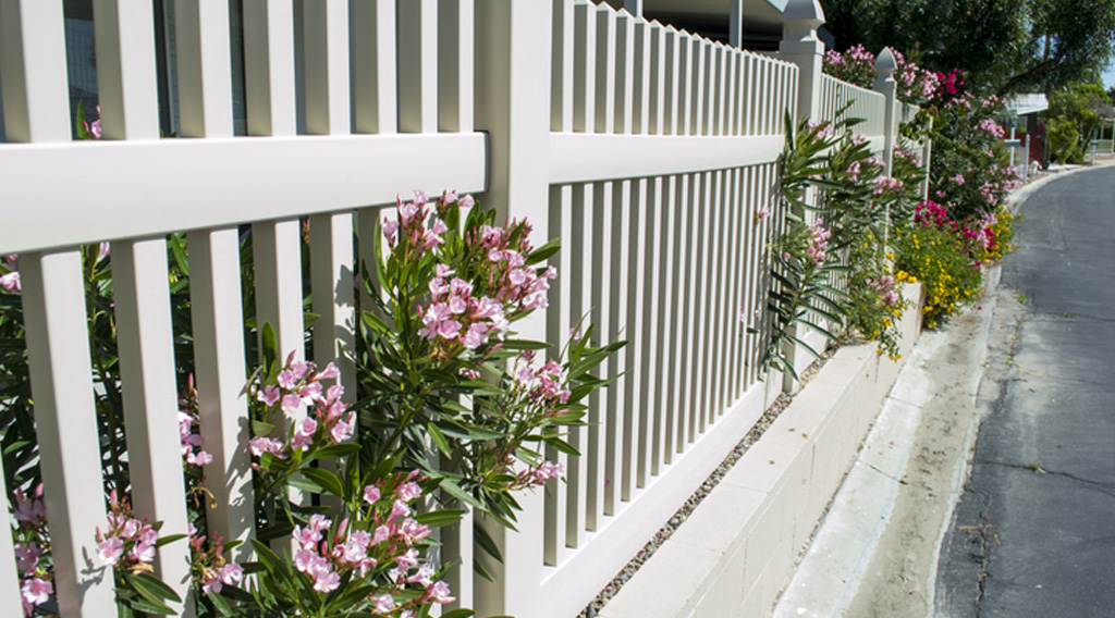 White vinyl fence with plants poking through over sidewalk