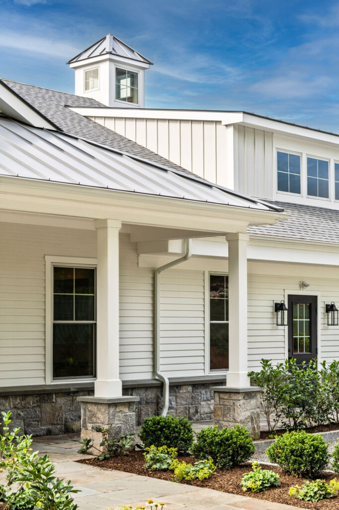 A modern looking guest house off to the side of a main house featuring immaculate landscaping and white siding atop a rock veneer footer.
