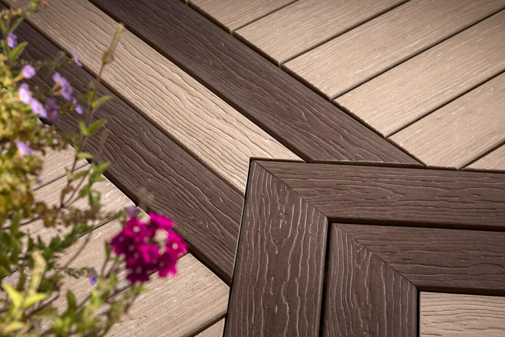 Intersecting light and dark brown decking boards at a corner with an out of focus plant to the left of the photo.