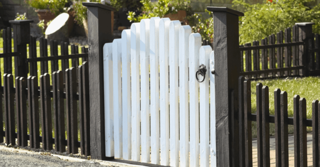 A dark brown lumber picket fence white a white gate door and ornamental handle.
