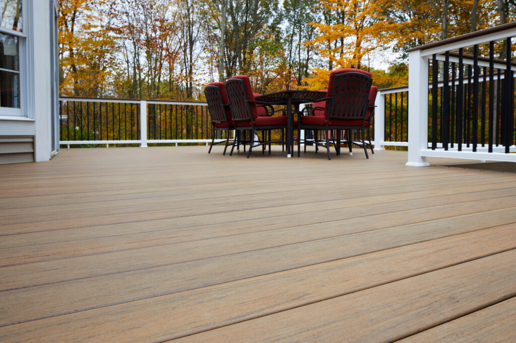 Down low to a dark brown deck with white railings, there is an outdoor table with red accent cushions in the corner.