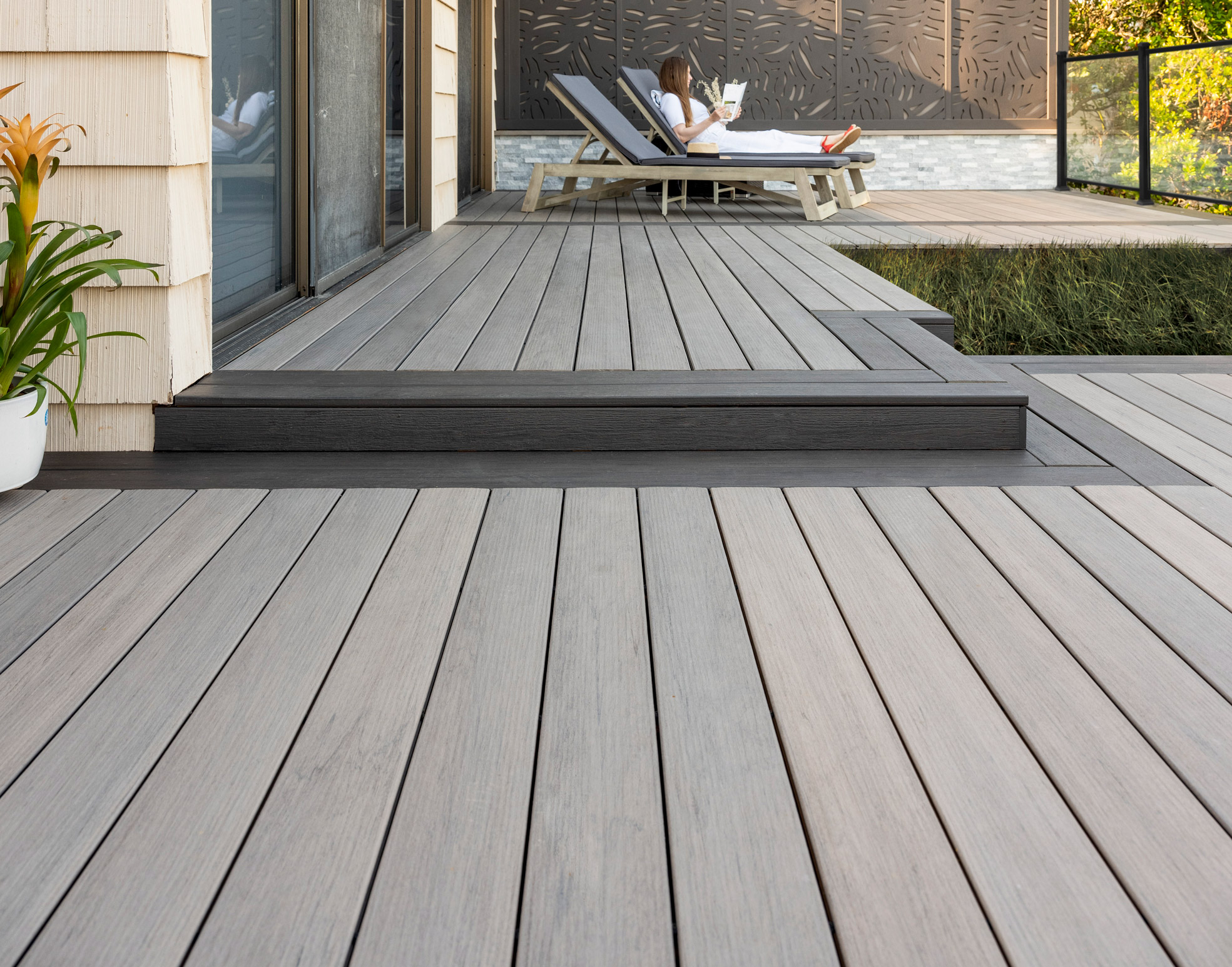 A close up shot of a modern looking deck featuring light gray decking bordered with dark gray accents, and two lounge chairs (one with a woman sitting on it reading a book) in the background.