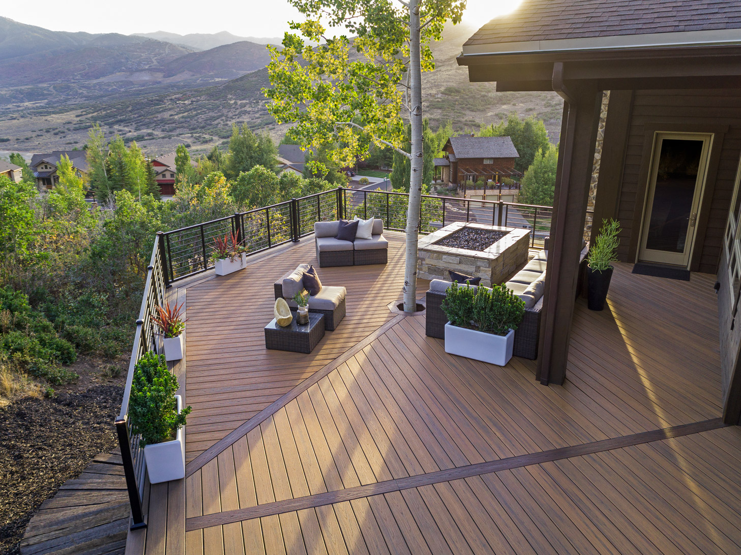 Looking down on a modern deck featuring a fire pit, potted plants, a central tree coming through the deck, and a a large seating area.